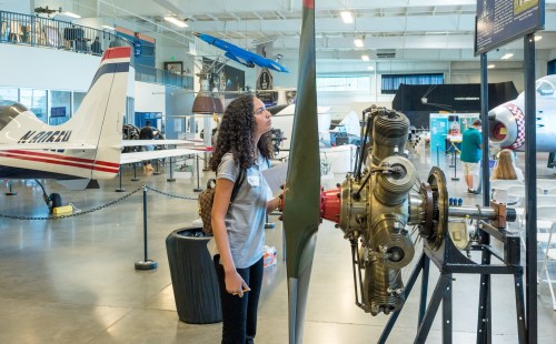 A woman examines an aircraft engine in an aviation museum, surrounded by various planes and exhibits.