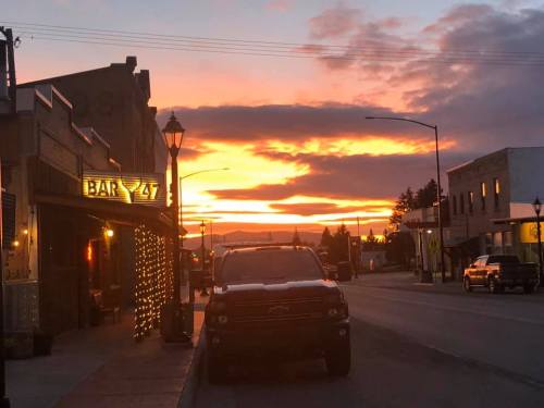 A vibrant sunset casts orange and pink hues over a small town street, with a bar and parked truck in view.