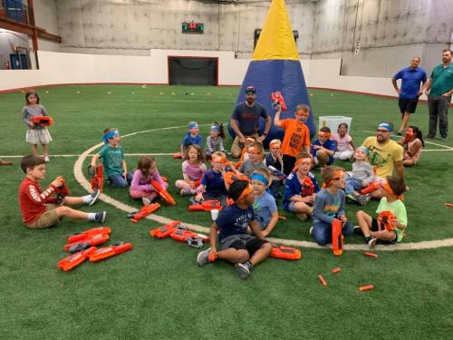 A group of children and adults in a sports facility, wearing blue headbands and holding toy blasters, gathered for a game.