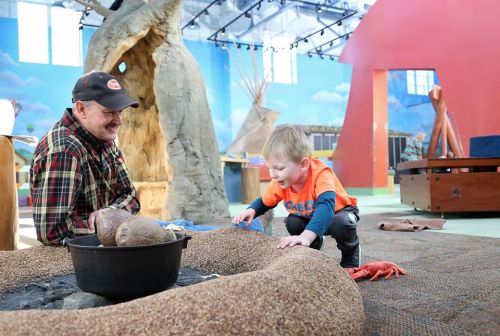 A man and a young boy play together on a textured surface in a colorful indoor space, surrounded by natural elements.