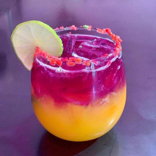 A colorful layered drink in a glass, featuring purple and orange hues, garnished with a lime slice and colorful rim.