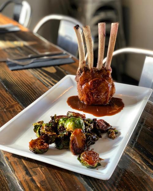 A plate featuring a rack of lamb with a rich sauce, accompanied by roasted Brussels sprouts and potatoes on a wooden table.