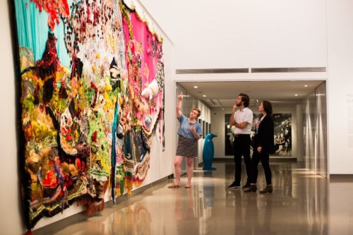 Visitors admire a vibrant textile artwork in an art gallery, discussing its details and colors.