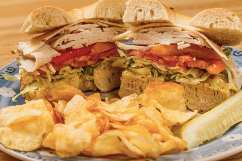 A close-up of a sandwich with turkey, cheese, lettuce, and tomato, served with potato chips and a pickle on the side.