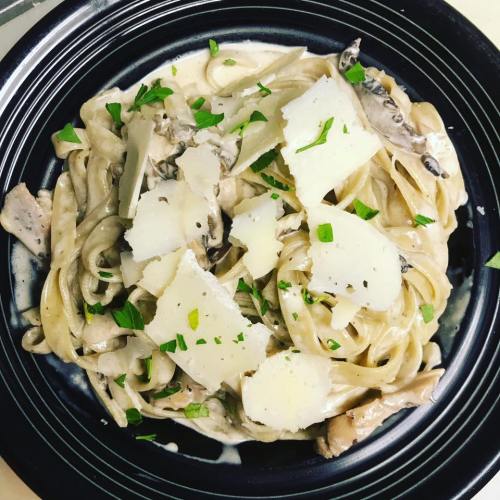 A plate of creamy fettuccine pasta topped with chicken, mushrooms, and shaved Parmesan, garnished with fresh herbs.