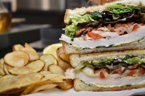 A close-up of a stacked sandwich with turkey, bacon, lettuce, and tomato, served with crispy potato chips on the side.