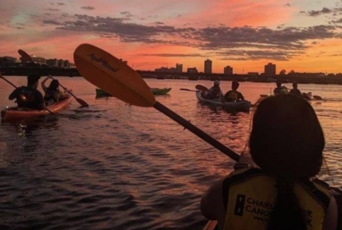 A group of kayakers paddles on a calm river at sunset, with city skyline silhouettes in the background.