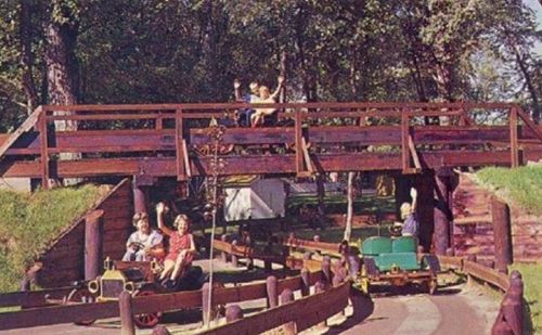 Two children wave from a wooden bridge above a small amusement park ride with colorful cars below.