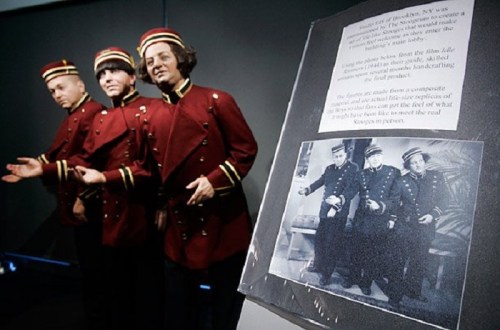 Three wax figures of bellhops in red uniforms stand beside a framed black-and-white photo of two men in similar attire.