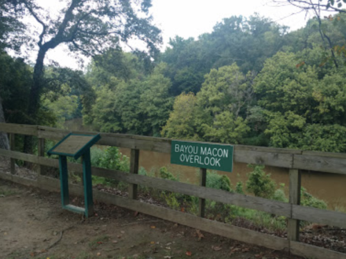 Sign for Bayou Macon Overlook beside a wooden fence, with a view of a river and lush green trees in the background.