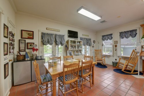 A cozy kitchen with a wooden dining table, rocking chairs, and checkered curtains, featuring a fridge and decorative items.