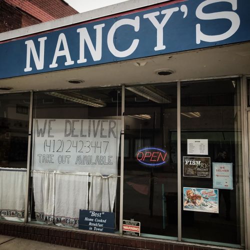 A storefront with "Nancy's" sign, delivery information, and an "Open" sign in the window. White curtains are visible.