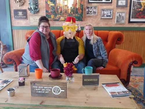 Two women sit on a couch with a person in a pig mask, all smiling in a cozy café setting. A "Reserved" sign is on the table.