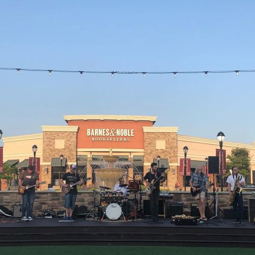 A band performs on an outdoor stage in front of a Barnes & Noble store, with string lights overhead and a fountain nearby.
