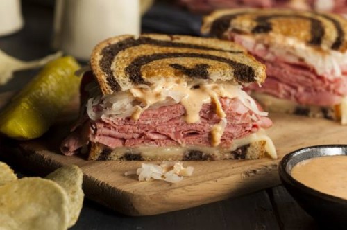 A close-up of a sliced sandwich with layers of meat, sauerkraut, and sauce on marbled rye bread, served with pickles and chips.