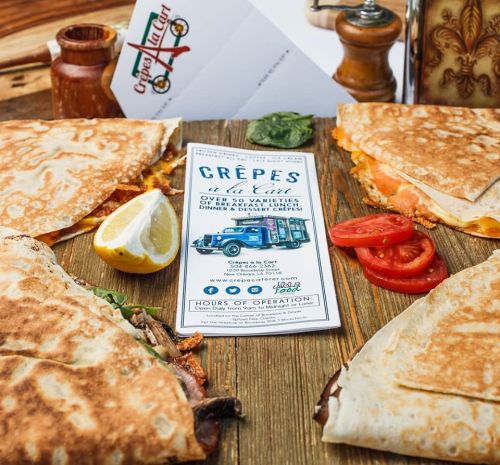 A wooden table with crepes, lemon, tomatoes, and a menu for a crepe restaurant featuring various options.