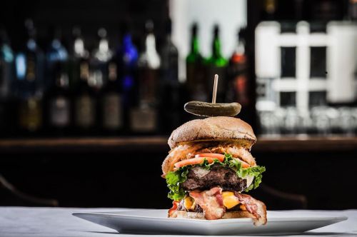 A towering burger with bacon, cheese, lettuce, tomato, and a pickle on top, served on a white plate.