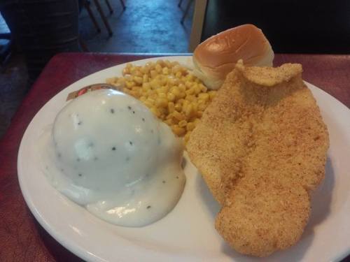 A plate with fried fish, creamy mashed potatoes with gravy, corn, and a soft dinner roll.