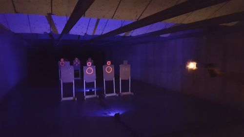 A dimly lit shooting range with targets illuminated by blue light, as a gun is fired towards them.