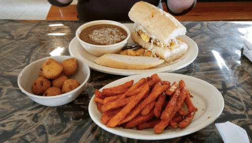 A plate with a sandwich, sweet potato fries, and a bowl of beans, alongside a dish of corn fritters.
