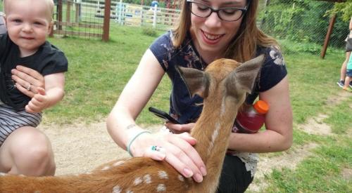 A woman smiles as she pets a young deer, while a baby sits beside her, both enjoying a sunny outdoor setting.