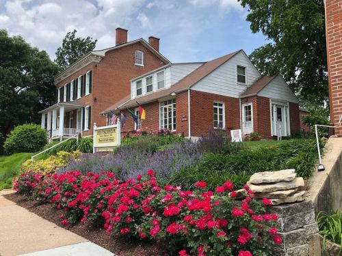 A brick building with white accents, surrounded by colorful flowers and flags, under a blue sky with clouds.