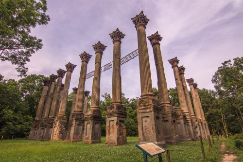 Ruins of tall, ornate columns in a grassy area, surrounded by trees under a cloudy sky. Historical site marker visible.