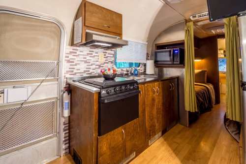 Interior of a vintage Airstream kitchen with wooden cabinets, a stove, microwave, and cozy decor.