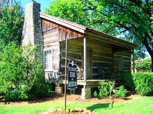 A rustic log cabin with a stone chimney, surrounded by greenery, featuring a sign for the "McGavock Home and Museum."