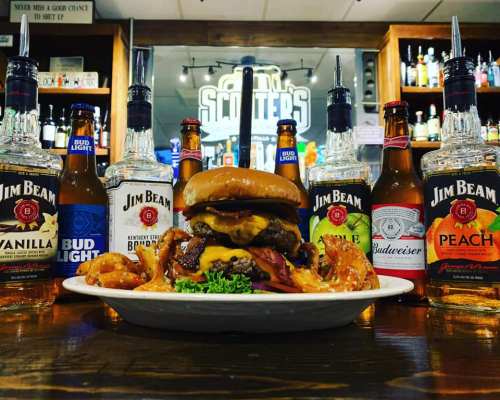 A juicy cheeseburger with bacon and fries, surrounded by various bottles of Jim Beam and Budweiser at a bar.