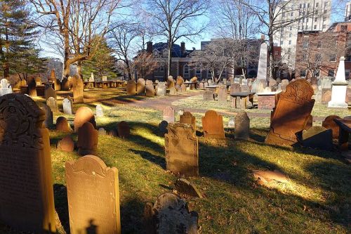 A peaceful cemetery with various gravestones, surrounded by trees and historic buildings in the background.