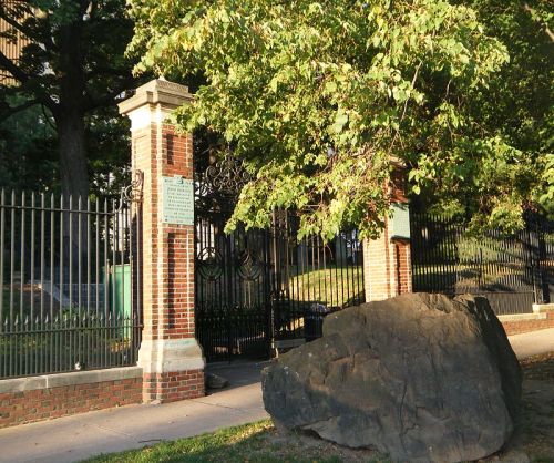 Entrance gate with brick pillars and wrought iron, surrounded by trees and a large rock in the foreground.