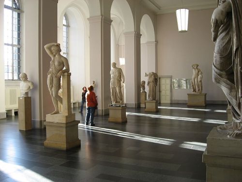 A spacious museum gallery featuring classical sculptures and two visitors admiring the artwork.