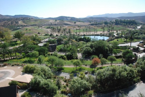 Aerial view of a lush park with trees, a pond, and rolling hills in the background under a clear blue sky.