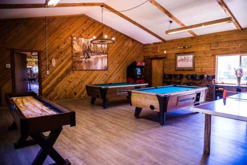 A cozy game room with wooden walls, featuring pool tables, a foosball table, and an arcade machine.