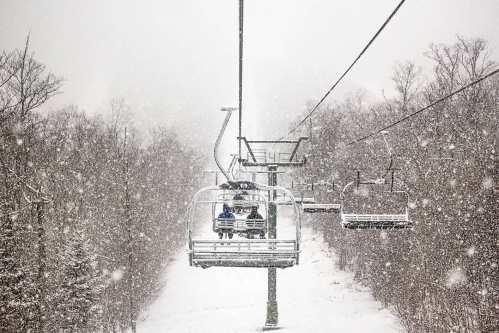 Two people sit on a ski lift surrounded by heavy snowfall and trees in a winter landscape.