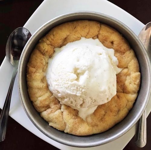 A warm cookie bowl topped with a scoop of vanilla ice cream, served in a metal dish on a white plate.
