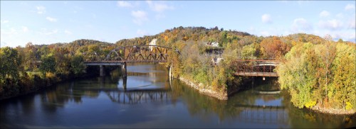 A panoramic view of two bridges over a calm river, surrounded by colorful autumn foliage and rolling hills.