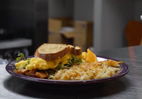 A plate of scrambled eggs, hash browns, bacon, and toast, garnished with a slice of orange.