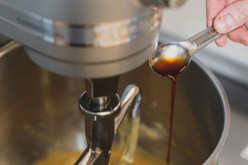 A hand holds a spoonful of vanilla extract above a mixing bowl with a stand mixer in the background.