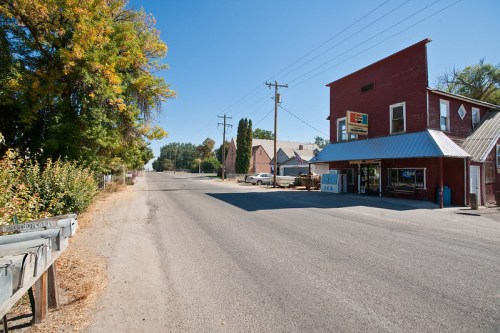 A quiet rural street with a red building, power lines, and trees lining the road. A small store is visible on the corner.