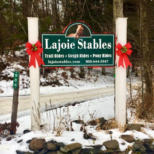 Sign for Lajoie Stables featuring trail rides, sleigh rides, and pony rides, decorated with red bows in a snowy setting.