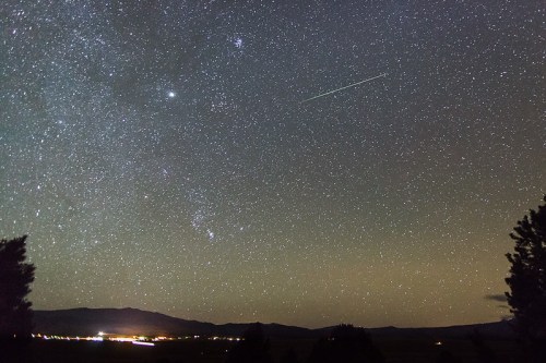 A starry night sky filled with stars, a bright meteor streaking across, and distant lights from a valley below.