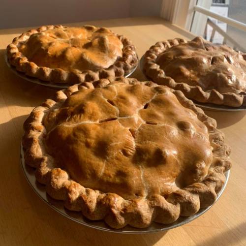 Three golden-brown pies with flaky crusts, resting on plates on a wooden table near a window.