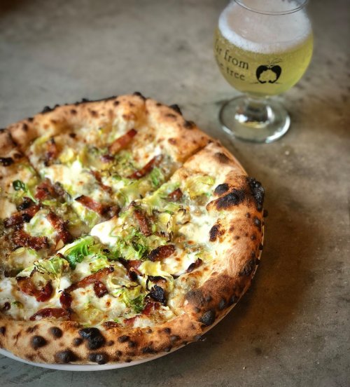 A freshly baked pizza topped with cheese, brussels sprouts, and sausage, alongside a glass of light beer.