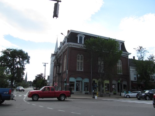 A red pickup truck turns at a street corner near a historic brick building with large windows and a church steeple in the background.
