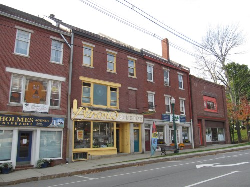 A row of brick buildings with various storefronts, including a studio, insurance agency, and a colorful sign.