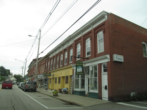 A street view of a small town featuring brick buildings, a pharmacy, and shops with power lines overhead.