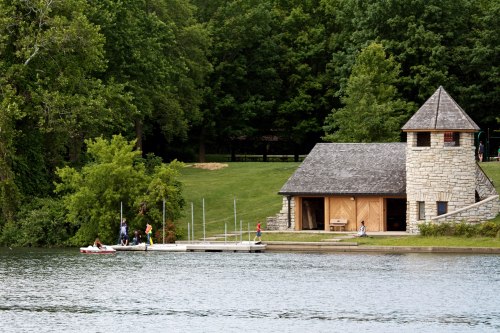 A serene lake scene featuring a stone building, dock, and people enjoying activities near the water surrounded by greenery.
