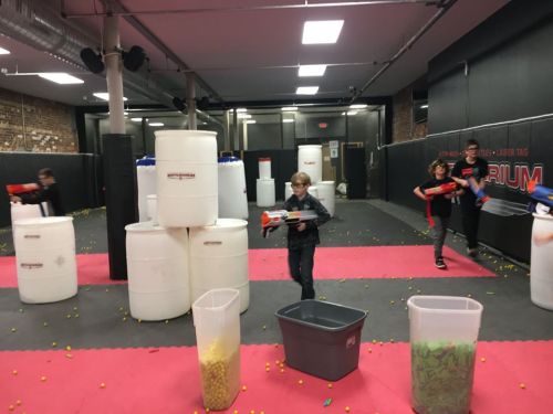 Kids playing laser tag in an indoor arena with obstacles and colorful foam balls scattered on the floor.